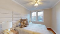 This single-floor residence offers a thoughtfully designed living environment with a refined aesthetic throughout. The bedroom is a standout feature, showcasing elegant crown molding that frames the ceiling and adds a touch of classic architectural character to the space. A ceiling fan with integrated lighting sits centrally overhead, providing both comfort and ambient illumination.

The room is bathed in natural light streaming through a pair of double-hung windows, each framed with clean white trim that complements the neutral, warm-toned walls. The windows offer views of the surrounding neighborhood, adding a sense of connection to the exterior environment.

A large upholstered headboard in a soft, cream fabric serves as the focal point of the bedroom, lending a sophisticated and contemporary feel. The bedding is kept in muted, neutral tones, accented by decorative pillows featuring geometric patterns in warm earth tones — a nod to modern interior styling. A compact nightstand with a simple table lamp rounds out the furnishings, contributing to the room's calm and restful atmosphere.

Overall, the space conveys a clean, comfortable, and well-maintained environment, blending classic architectural details with modern interior design sensibilities to create an inviting and functional living space.