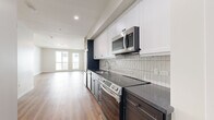 This thoughtfully designed single-floor unit offers an efficient and modern living experience. The open-plan layout seamlessly connects the kitchen and living area, creating a cohesive and functional space bathed in natural light from a pair of large windows that overlook an exterior balcony railing.

The kitchen is a standout feature, boasting a sleek two-tone design with crisp white upper cabinetry and dark charcoal lower cabinets topped with dark stone countertops. A vertically laid white tile backsplash adds texture and visual interest. Stainless steel appliances — including a refrigerator, dishwasher, electric range, and over-the-range microwave — complete the polished, contemporary look. Warm hardwood-style flooring runs continuously throughout the living area and kitchen, grounding the space with a welcoming tone.

The bathroom features classic black-and-white hexagonal mosaic tile flooring, a clean white vanity with a single sink, a large mirror, and a full tub-shower combination enclosed by a glass panel, giving the space a bright and timeless character.

Two closets provide practical storage solutions throughout the unit. Crisp white walls, recessed lighting, and clean architectural lines contribute to an overall ambiance that feels modern, bright, and move-in ready.