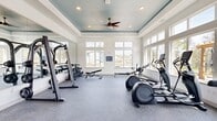 Located at 500 River Knoll Way in Columbus, Georgia, this fitness center offers a thoughtfully designed workout environment that combines functionality with refined architectural details. The space is defined by a distinctive tray ceiling painted in a deep teal-blue hue, accented by recessed lighting and a ceiling fan that keeps the atmosphere comfortable during intense workouts. Large, multi-pane windows line the walls on multiple sides, flooding the interior with natural light and offering views of the surrounding outdoor amenity area, including a pergola, pool, and lush tree line. The durable rubber-speckled flooring provides both traction and cushioning underfoot, making it well-suited for a variety of exercises. A full-length wall mirror expands the visual footprint of the room and is complemented by a mounted flat-screen television, adding an entertainment element to the workout experience. The equipment selection is comprehensive, including treadmills, elliptical trainers, a stationary bike, a rowing machine, a multi-function cable machine, and a tiered dumbbell rack paired with a flat bench for strength training. The overall ambiance is bright, clean, and welcoming, striking a balance between a professional gym atmosphere and the comfort of a community fitness facility.