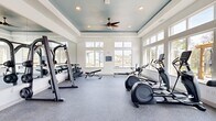 Located at 500 River Knoll Way in Columbus, Georgia, this fitness center offers a thoughtfully designed workout environment that combines functionality with refined architectural details. The space is defined by a distinctive tray ceiling painted in a deep teal-blue hue, accented by recessed lighting and a ceiling fan that keeps the atmosphere comfortable during intense workouts. Large, multi-pane windows line the walls on multiple sides, flooding the interior with natural light and offering views of the surrounding outdoor amenity area, including a pergola, pool, and lush tree line. The durable rubber-speckled flooring provides both traction and cushioning underfoot, making it well-suited for a variety of exercises. A full-length wall mirror expands the visual footprint of the room and is complemented by a mounted flat-screen television, adding an entertainment element to the workout experience. The equipment selection is comprehensive, including treadmills, elliptical trainers, a stationary bike, a rowing machine, a multi-function cable machine, and a tiered dumbbell rack paired with a flat bench for strength training. The overall ambiance is bright, clean, and welcoming, striking a balance between a professional gym atmosphere and the comfort of a community fitness facility.