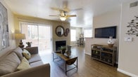 This single-floor space offers a well-appointed, open-concept layout designed for comfortable everyday living. The main living area is anchored by a tile-surround fireplace, which serves as a natural focal point and adds warmth to the room. Hardwood-style flooring runs throughout, lending a clean and cohesive aesthetic to the space. A ceiling fan with an integrated light fixture keeps the area comfortable and well-lit. Large sliding glass doors with vertical blinds open to an outdoor patio, filling the room with natural light and providing easy access to the exterior. Multiple windows further enhance the bright and airy atmosphere. The living area is generously sized, offering ample space for seating and entertainment, complemented by a wall-mounted television setup and a rustic open shelving unit. Decorative wall accents, including circular mirrors and gear-shaped ornaments, add a distinctive, personalized character to the neutral-toned walls. A bar-height dining area is visible in the background, creating a natural transition between the living and dining spaces. The textured ceiling adds architectural depth throughout the interior, completing the overall inviting and functional character of this home.
