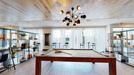Located at 444 W College Ave in Tallahassee, Florida, this single-floor community amenity space offers a generously sized, thoughtfully designed environment for recreation and socializing. The interior is unified by warm light wood plank ceilings, light hardwood-style flooring, and crisp white walls that create a bright, contemporary atmosphere throughout.

The space is divided into two distinct zones. The game room area is a standout, featuring a full-size billiards table as its centerpiece, complemented by a ping pong table, foosball table, air hockey table, and a large-format connect-four wall game. A wall-mounted flat-screen TV is framed by a wood accent wall, and plush leather seating provides comfortable viewing. A striking mid-century modern chandelier with spherical brass pendants anchors the room visually.

The lounge area exudes a relaxed, social ambiance with oversized sectional sofas, eclectic accent chairs, and curated wall art. A sleek open shelving unit with decorative accessories adds character and visual depth. A distinctive wet bar or service counter features mint-green cabinetry, a dramatic marble backsplash, and soft seafoam tile above, offering an eye-catching design moment. Floor-to-ceiling sheer curtains line the exterior wall, allowing ample natural light while maintaining an airy, sophisticated feel.