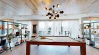 Located at 444 W College Ave in Tallahassee, Florida, this single-floor community amenity space offers a generously sized, thoughtfully designed environment for recreation and socializing. The interior is unified by warm light wood plank ceilings, light hardwood-style flooring, and crisp white walls that create a bright, contemporary atmosphere throughout.

The space is divided into two distinct zones. The game room area is a standout, featuring a full-size billiards table as its centerpiece, complemented by a ping pong table, foosball table, air hockey table, and a large-format connect-four wall game. A wall-mounted flat-screen TV is framed by a wood accent wall, and plush leather seating provides comfortable viewing. A striking mid-century modern chandelier with spherical brass pendants anchors the room visually.

The lounge area exudes a relaxed, social ambiance with oversized sectional sofas, eclectic accent chairs, and curated wall art. A sleek open shelving unit with decorative accessories adds character and visual depth. A distinctive wet bar or service counter features mint-green cabinetry, a dramatic marble backsplash, and soft seafoam tile above, offering an eye-catching design moment. Floor-to-ceiling sheer curtains line the exterior wall, allowing ample natural light while maintaining an airy, sophisticated feel.