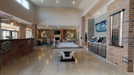 Located at 600 67th Ave N in Myrtle Beach, South Carolina, this single-floor community clubhouse is thoughtfully designed to serve as a social and recreational hub. The open-concept layout is anchored by soaring vaulted ceilings and exposed brick columns that lend an industrial-chic character throughout the space, while polished concrete flooring unifies the main gathering areas.

A central lounge area features a curved gray sectional sofa set atop a vibrant teal carpet, complemented by coastal-themed artwork, decorative pendant lighting, and a brick-surround fireplace with a wall-mounted television above. The seating arrangement is inviting and well-suited for both casual gatherings and community events.

Adjacent to the lounge, a billiards table occupies a prominent position beneath recessed lighting, with additional seating areas nearby. A dedicated refreshment station includes built-in cabinetry, a coffee bar, and stainless steel appliances.

The bar area is a standout feature, boasting a full-length granite countertop supported by a brick base and lined with modern bar stools. Large overhead garage-style doors open to allow natural light and outdoor views, creating a seamless indoor-outdoor connection. The overall ambiance balances coastal warmth with modern amenities throughout.