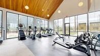 This contemporary fitness center is defined by its striking architectural design, most notably the dramatic vaulted ceiling clad in warm wood paneling that soars overhead, creating an airy and expansive atmosphere. Pendant globe lights hang at varying heights throughout, complementing the abundant natural light that pours in through the expansive floor-to-ceiling windows lining the perimeter. These large-format windows frame views of the surrounding landscape, including greenery, trees, and the broader neighborhood beyond.

The facility is organized across several interconnected zones, each dedicated to a different aspect of fitness. The main cardio area features a full lineup of treadmills, ellipticals, stair climbers, and stationary bikes equipped with individual screens. A dedicated cycling studio houses additional spin bikes in a space framed by warm wood-paneled walls and large windows. The strength training zone offers a robust Torque multi-functional rack, free weights, benches, and cable machines, while a functional training area provides TRX suspension trainers, kettlebells, medicine balls, BOSU trainers, and foam rollers neatly organized on a storage rack. Glass partition walls and doors divide the zones while maintaining an open, connected feel. Large-format tile and polished concrete flooring complete the clean, modern aesthetic throughout.