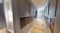 This sophisticated mail room and corridor space exemplifies refined residential common area design. The hallway is lined on both sides with numbered bronze U.S. mailboxes, accommodating a large number of residents and organized in clearly labeled rows. The mailboxes are set within paneled wainscoting finished in soft grey tones, lending a polished, boutique-hotel quality to what is typically a purely functional space.

The floor is a standout architectural feature — a light natural wood parquet laid in an intricate geometric star-and-diamond pattern that draws the eye down the length of the corridor. Crown molding and recessed lighting overhead add to the sense of elegance and height.

At the far end of the mail corridor, a decorative console table with a framed mirror and a brass-accented cabinet provides a refined finishing touch. Wall-mounted sconces in a brushed gold finish contribute warm accent lighting along the passage.

The corridor opens into a broader common area, where the same striking parquet flooring continues, leading into a furnished lounge with seating, decorative partitions with gold detailing, and additional gathering spaces. The overall ambiance is one of upscale, thoughtfully curated design that balances practicality with aesthetic sophistication throughout every element of the shared space.
