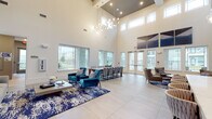 This elegantly designed single-floor amenity space makes an immediate impression with its dramatic double-height ceilings that lend an expansive, airy quality throughout. Large windows line the walls at multiple levels, flooding the interior with generous natural light while offering views of the surrounding community and outdoor areas.

The open-plan layout is thoughtfully organized into distinct functional zones. A generous lounge area anchors the space with plush, jewel-toned velvet seating in vibrant teal and complementary neutral tones, arranged atop richly patterned navy and ivory area rugs over large-format light tile flooring. A sculptural globe chandelier suspended from the soaring ceiling adds a refined focal point overhead.

A standout wet bar area features a striking mosaic metallic tile backsplash, a marble-topped counter with stylish woven bar stools, and pendant lighting — creating a sophisticated social hub within the space. Adjacent to the lounge, a long communal dining table with a sleek marble surface and modern upholstered chairs provides an ideal setting for group gatherings, with direct access to an outdoor area through glass doors.

Additional seating vignettes with floor lamps, side tables, and curated décor contribute to the space's warm yet contemporary ambiance. The overall aesthetic is modern and polished, combining clean architectural lines with carefully selected furnishings and finishes.