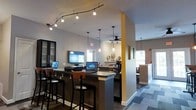Located at 300 Autumn River Run in the Roxborough neighborhood of Philadelphia, this single-floor community clubhouse offers a thoughtfully designed space that balances functionality with a welcoming ambiance. The business center area features a sleek, dark-toned counter bar equipped with workstations and laptops, illuminated by a striking curved track lighting fixture overhead, providing ample task lighting for residents working or studying. Pendant lights with an industrial flair hang throughout, adding visual interest and warmth to the space. A bold yellow structural column serves as a design accent, punctuating the open floor plan and delineating the various zones within the clubhouse. A ceiling fan ensures comfortable airflow in the adjoining lounge area, which benefits from natural light streaming through multiple windows. A wall-mounted flat-screen display is positioned for easy viewing from the workstation bar. Toward the back, a café-style setup with a coffee station and seating area provides a comfortable spot for casual gatherings. The flooring transitions between tile and carpet, further defining the distinct functional zones. Overall, the space delivers a clean, modern aesthetic with practical amenities tailored to an active residential community.
