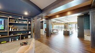 Located on Indian Creek Road NW in Huntsville, Alabama, this single-floor community clubhouse is designed with a contemporary aesthetic that blends warm wood tones, bold accent colors, and thoughtfully curated furnishings throughout its open, flowing layout.

The main gathering area features wood-look tile flooring, substantial wood-clad structural columns, and a coffered ceiling with exposed beam detailing finished in rich walnut tones. A built-in shelving unit with a deep navy backdrop anchors one end of the space, displaying decorative objects alongside an embedded flat-screen television, creating an inviting lounge atmosphere with comfortable seating nearby.

At the heart of the space sits a striking curved kitchen island finished in sage green with a crisp white countertop, surrounded by mustard-yellow bar stools. Behind it, brass-framed open shelving reflects light against an antiqued mirror backdrop, complemented by a full run of cabinetry. Large floor-to-ceiling windows and glass doors frame views of an outdoor pool area, flooding the interior with natural light.

A separate lounge zone features blue accent walls, exposed wood beam ceilings, mid-century inspired furnishings, and a wall-mounted television with generous window views. Restroom facilities and additional seating nooks complete the layout, making this clubhouse a well-appointed, multifunctional communal space.
