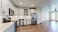 This single-floor residence offers a clean, contemporary design throughout, combining functional layout with modern finishes. The open-concept living and kitchen area serves as the heart of the space, with warm hardwood-style flooring extending across the main living zone and recessed lighting overhead that creates a bright, welcoming atmosphere.

The kitchen is thoughtfully appointed with crisp white shaker-style cabinetry that extends to the ceiling, providing generous storage. Stainless steel appliances — including a side-by-side refrigerator with ice and water dispenser, a dishwasher, and a range with microwave — offer a polished, cohesive look. White tile backsplash and light countertops complete the clean aesthetic, while open shelving adds a touch of character.

The bedroom features plush carpeting and connects directly to a well-designed en-suite bathroom. The bathroom is finished with large-format tile flooring, a single vanity with light wood cabinetry, and a separate glass-enclosed walk-in shower accented with chrome fixtures. An adjacent walk-in closet provides ample storage with wire shelving and hanging rods.

Throughout the space, neutral tones, white panel doors, and consistent finishes create a cohesive and move-in-ready environment that balances comfort with modern style.