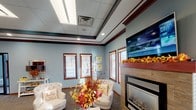 This single-floor space is designed to create a warm and inviting atmosphere, with thoughtful architectural details that enhance its overall character. The most notable room visible is a comfortable lounge or waiting area, finished with soft blue walls that lend a calm, welcoming tone throughout. A tray ceiling with rich dark wood trim adds a layer of architectural refinement, drawing the eye upward and giving the room a sense of defined elegance. Recessed lighting and flush-mounted ceiling fixtures ensure the space is well-lit, complementing the natural light that filters in through multiple windows dressed with white horizontal blinds and framed in warm-toned wood trim. A tile-surround fireplace serves as a central focal point along one wall, topped by a wall-mounted flat-screen television, making the area both functional and comfortable. Plush accent chairs with nailhead detailing provide seating, while seasonal floral arrangements add decorative charm. The combination of carpeted flooring, decorative crown molding, and carefully chosen furnishings contributes to a polished yet approachable ambiance. Overall, the space balances professional presentation with a hospitable, comfortable feel suited for welcoming guests or clients.