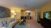 This single-floor residence offers an open-concept layout that effortlessly blends the living and dining areas into one cohesive, comfortable space. The living area is anchored by a plush, neutral-toned sofa accented with decorative pillows, a stylish geometric coffee table, and a classic tripod floor lamp that casts a warm, ambient glow throughout the room. A decorative ladder shelf adds a contemporary touch to the corner, complementing the overall aesthetic. The dining area flows naturally from the living space and features a dark rectangular dining table surrounded by upholstered grey chairs, illuminated by a sleek track lighting fixture overhead. A framed piece of wall art serves as a focal point behind the dining table, adding visual interest and depth. Soft, neutral carpeting runs throughout both areas, contributing to the warm and inviting ambiance. The light-toned walls and consistent flooring create a sense of openness and continuity, making the space feel expansive and well-connected. The layout is both functional and aesthetically considered, offering a comfortable environment suited for everyday living and casual entertaining alike.