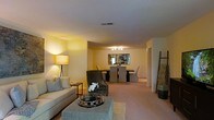 This single-floor residence offers an open-concept layout that effortlessly blends the living and dining areas into one cohesive, comfortable space. The living area is anchored by a plush, neutral-toned sofa accented with decorative pillows, a stylish geometric coffee table, and a classic tripod floor lamp that casts a warm, ambient glow throughout the room. A decorative ladder shelf adds a contemporary touch to the corner, complementing the overall aesthetic. The dining area flows naturally from the living space and features a dark rectangular dining table surrounded by upholstered grey chairs, illuminated by a sleek track lighting fixture overhead. A framed piece of wall art serves as a focal point behind the dining table, adding visual interest and depth. Soft, neutral carpeting runs throughout both areas, contributing to the warm and inviting ambiance. The light-toned walls and consistent flooring create a sense of openness and continuity, making the space feel expansive and well-connected. The layout is both functional and aesthetically considered, offering a comfortable environment suited for everyday living and casual entertaining alike.