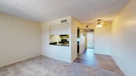 This single-floor residence offers a straightforward and functional layout designed for comfortable everyday living. The living area is finished with plush carpeting and features a textured ceiling, a ceiling fan with light fixture, and a pass-through window that connects directly to the kitchen—creating an open, communicative flow between the two spaces. The kitchen is equipped with dark granite-look countertops, warm wood-toned cabinetry, a black electric coil range with overhead exhaust hood, and wood-look vinyl flooring, providing a practical and efficient cooking environment. A dedicated dining or transitional space connects the kitchen to the rest of the unit, also finished with wood-look flooring for easy maintenance. The bedroom is generously sized with soft carpeting, neutral walls, and vertical blinds covering a window or sliding door that allows natural light to filter in. The bathroom is clean and functional, featuring a full tub and shower combo with a white surround, wood-look flooring, and chrome fixtures and towel bars. Throughout the unit, warm neutral tones and consistent finishes create a cohesive and livable ambiance suited for a range of occupants.
