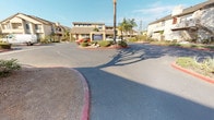 This residential community makes a strong first impression with a clearly marked entrance featuring a prominent "Welcome" sign and a detailed community map for easy navigation. Tall palm trees and mature shade trees are thoughtfully distributed throughout the property, lending a warm, established character to the grounds. The entry area is flanked by neatly trimmed shrubs, decorative boulders, and drought-tolerant plantings set against red-curbed landscaped medians, reflecting a cohesive and well-maintained aesthetic. The multi-story residential buildings feature tan and brown exterior finishes with pitched rooflines, private balconies, and exterior staircases providing access to upper levels. A controlled-access gate with metal fencing secures the interior of the community, offering residents a sense of privacy and security. Interior roadways wind through the property, lined with additional mature trees and low-growing plants that provide shade and visual appeal. Metal fencing borders portions of the community perimeter, further defining the boundaries of the space. The overall ambiance is that of a peaceful, established residential setting with a strong emphasis on landscaping, accessibility, and community organization, all enjoyed under an abundance of natural sunlight.