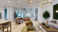 Located in Miramar, Florida, this single-story community clubhouse at 4260 SW 117th Ln is a thoughtfully designed gathering space that blends modern sophistication with a warm, resort-inspired atmosphere. Light-toned wood-look tile flooring flows seamlessly throughout, unifying the open layout and reflecting the abundant natural light that streams in through large black-framed windows and glass doors.

The entrance greets visitors with a striking round wooden reception table adorned with a fresh floral arrangement, setting an inviting tone. A decorative ceiling with a geometric gold-patterned inlay adds a luxurious first impression. Beyond the foyer, the main lounge opens into a generously proportioned gathering area featuring coffered ceilings with exposed wooden beams, elegant chandelier lighting, and multiple seating arrangements designed for socializing and relaxation.

A standout feature wall clad in marble-veined stone anchors a large flat-screen display, flanked by vertical slat paneling for a refined architectural accent. A bar-height seating area with warm wood cabinetry and pendant lighting offers a casual hub near windows overlooking lush outdoor scenery, including views of palm trees and the pool area.

A dedicated game room with a billiards table and large sectional seating provides recreational space, while a glass-walled private conference room with gold lantern pendants serves more formal needs. Throughout, curated artwork, indoor greenery, and gold-accented fixtures contribute to the space's polished, contemporary character.