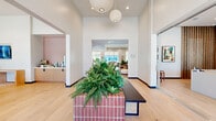 This thoughtfully designed single-floor amenity space offers a diverse range of functional areas connected by a cohesive modern aesthetic. The grand entry welcomes visitors with soaring ceilings, warm light wood flooring, and a striking tiled planter bench adorned with lush greenery, anchored by elegant globe pendant lighting overhead. Adjacent to the entry, a large open lounge area features polished concrete floors, a large-screen display, wood slat room dividers, and an eclectic mix of seating arrangements suited for both relaxation and socializing. The fully equipped kitchen and bar area is a standout feature, showcasing a spacious island with terracotta tile accents, light wood cabinetry, and a wall-mounted TV — ideal for community gatherings. A dedicated dining area with natural wood tables, velvet upholstered chairs, and a circular pendant light provides a warm, communal setting. Private meeting rooms offer intimate round tables, plush upholstered seating, patterned area rugs, and soft diffused natural light through shaded windows. A co-working nook provides a built-in wood desk surface, acoustic wall paneling, and views to the outdoors. Throughout the space, curated artwork, indoor plants, and layered textures contribute to a refined yet welcoming atmosphere.
