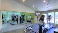 This fitness center offers a comprehensive workout environment designed to accommodate a range of exercise routines. The space is finished with durable speckled flooring suited for high-traffic athletic use, while vaulted ceilings contribute to an open, airy atmosphere throughout.

The cardio area is well-stocked with equipment including a stationary bike, an elliptical trainer, and additional cardio machines positioned to take advantage of natural light from a window along the far wall. A wall-mounted flat-screen TV is positioned at a comfortable viewing angle, allowing members to stay entertained during their workouts.

Along one wall, floor-to-ceiling mirrors visually expand the space and allow users to monitor their form during exercises. The strength training section features a multi-use cable and weight machine, along with wall-mounted accessory stations, providing options for a full-body resistance workout.

The walls are painted in a clean combination of white and vibrant green, lending the space an energetic and motivating ambiance. Recessed lighting throughout ensures the entire area is well-lit. Informational boards are mounted on the wall, offering workout guidance and resources to users. Overall, the fitness center is thoughtfully arranged to maximize functionality and encourage an active lifestyle.