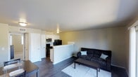 This single-floor residence offers a practical and modern open-concept layout that seamlessly connects the living area and kitchen. The living room is comfortably sized, featuring dark hardwood-style flooring that extends throughout the main living space, a neutral-toned area rug, and smooth light gray walls that create a clean, contemporary backdrop. A dark upholstered sofa paired with a rectangular coffee table anchors the living space, lending it a cozy yet functional feel.

The kitchen is separated from the living area by a low half-wall counter that serves as a natural divider while maintaining the open flow of the space. White cabinetry lines the kitchen walls, offering ample storage, while the countertop workspace provides a practical surface for meal preparation. Black appliances, including a gas range, microwave, and refrigerator, add a bold contrast to the white cabinetry. A ceiling-mounted light fixture provides central illumination, and a visible air vent suggests a built-in HVAC system. A white interior door near the entry area adds to the clean, cohesive aesthetic of the space. Overall, the residence presents a tidy and well-organized environment with a modern sensibility.