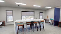 This contemporary single-floor workspace is thoughtfully designed to support both collaborative and focused work. The main area centers around a large, sleek white communal table flanked by colorful mid-century-style bar stools in vibrant teal, orange, and red tones, adding a lively visual contrast to the otherwise neutral, light-gray palette of the walls and carpeted flooring. Flush rectangular LED panel lighting overhead ensures consistent, bright illumination throughout the space. Multiple windows fitted with roller shades line the walls, allowing natural light to filter in while maintaining glare control and offering views of the surrounding outdoor environment. Along one side of the room, a dedicated workstation area features wood-toned desks with red accents, ergonomic white chairs, and desktop computers, partially separated by a bold blue dividing partition that adds a strong architectural accent. A built-in refreshment station with dark cabinetry, a countertop, and coffee-making appliances is tucked into one corner beneath pendant lighting, providing a convenient amenity. A cork bulletin board and additional wall-mounted accessories contribute functional touches throughout. The overall ambiance is clean, modern, and welcoming — well-suited for a business center, study lounge, or community amenity space.