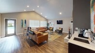 Located at 309 NE Fairgrounds Rd in Bremerton, Washington, this single-floor community clubhouse offers a thoughtfully designed gathering space that balances comfort and functionality. The centerpiece of the space is a spacious lounge area anchored by soaring vaulted ceilings with recessed lighting that floods the room with a bright, airy feel. Warm hardwood-style flooring runs throughout the main area, complementing the mid-century modern furnishings, including leather sofas, accent chairs, and bar-height seating. A white brick fireplace with a decorative mantle serves as a striking focal point, while a wall-mounted TV and curated artwork add character to the room. Large windows with blinds allow natural light to pour in, enhancing the inviting atmosphere. An adjacent kitchenette area features dark cabinetry, a countertop sink, and a coffee station, providing residents with convenient refreshment options. Vibrant, colorful artwork adorns the walls throughout, adding energy and personality to the space. The fitness center is well-appointed with treadmills, elliptical machines, and additional cardio equipment set on teal patterned carpet. Full-wall mirrors, ceiling fans, and multiple windows make the gym feel open and functional. Together, these spaces create a versatile and welcoming environment for community use.