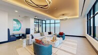 Located at 17071 W Dixie Hwy in North Miami Beach, FL, this elegantly designed lounge space offers a refined and contemporary atmosphere on a single floor. The living room serves as the centerpiece of the space, thoughtfully appointed with a curated mix of modern furnishings, including a generous white sofa, teal accent chairs, and a striking marble coffee table arrangement that anchors the room.

Overhead, a dramatic coffered ceiling with warm ambient cove lighting draws the eye upward, complemented by two sculptural, orbital pendant light fixtures that add an artistic focal point to the space. The clean white walls and light-toned large-format tile flooring create a bright, airy backdrop that enhances the overall sense of openness.

Floor-to-ceiling windows with black-framed curtain wall glazing flood the interior with abundant natural light and frame breathtaking views of the Miami skyline, a lush green roof terrace, and the shimmering waterway beyond. A private dining nook with navy upholstered booth seating and a high-gloss table offers an intimate setting within the space, while contemporary artwork adorning the walls adds a vibrant cultural touch. The seamless blend of natural light, sophisticated materials, and modern design makes this space a polished and inviting environment.
