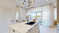 This thoughtfully designed single-floor residence showcases a cohesive modern aesthetic throughout, anchored by light hardwood flooring and a bright, neutral palette. The open-concept kitchen serves as the heart of the space, featuring a striking marble waterfall island topped with a matte black undermount sink and a sculptural branching chandelier with globe pendants overhead. Light wood cabinetry lines the walls alongside stainless steel appliances, including a gas range and French door refrigerator, while gold bar stools add a warm, contemporary accent.

Adjacent to the kitchen, the dining nook benefits from floor-to-ceiling glass doors that open to a private balcony, inviting an abundance of natural light and outdoor views. The living area offers a cozy yet refined atmosphere, centered around a linear electric fireplace with a wall-mounted television above and flanked by sleek vertical sconces.

The bedroom is generously lit by a large multi-pane picture window framing outdoor views, complemented by warm brass lighting fixtures and natural wood furnishings. The bathroom presents a spa-like finish with large-format stone tile surround, a soaking tub, black-framed glass partition, and polished gold fixtures throughout.