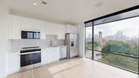 Located on the fifth floor at 616 W Fulton St in Grand Rapids, Michigan, this modern 2-bedroom, 1-bathroom unit offers a well-designed layout with contemporary finishes throughout. Light hardwood flooring runs continuously across the entire space, complementing the clean white walls and creating a bright, cohesive aesthetic.

The open kitchen is a standout feature, equipped with a full suite of stainless steel appliances including a French door refrigerator, electric range, built-in microwave, and dishwasher. Glossy white cabinetry extends to the ceiling, paired with a classic subway tile backsplash and ample counter space. A dramatic floor-to-ceiling window wall floods the kitchen and living area with natural light while framing impressive views of the Grand Rapids skyline and surrounding neighborhood.

Both bedrooms are well-proportioned and bright, each featuring large windows with city and neighborhood views. The primary bedroom includes a set of bold dark double doors, adding a modern architectural accent. The secondary bedroom is equally light-filled with its own oversized window.

The bathroom features a full tub-shower combo, a sleek floating vanity with white cabinetry, and coordinating light hardwood-style tile flooring. Throughout the unit, dark-framed windows and doors provide sharp contrast against the white interiors, reinforcing the space's modern, urban character.