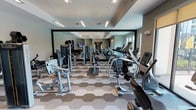 Located at 351 Northwood Dr in Flower Mound, Texas, this single-floor fitness center offers a well-appointed and thoughtfully designed space for a comprehensive workout experience. The main gym area is outfitted with a wide selection of cardio machines — including treadmills, ellipticals, and stationary bikes — alongside a robust lineup of strength and resistance training equipment. A distinctive hexagonal-patterned carpet tiles the floor, adding a modern geometric aesthetic throughout the space. Floor-to-ceiling mirrors along one wall amplify the sense of openness, while recessed and pendant lighting ensures the area is bright and functional. A built-in water station with a mosaic tile accent wall and a coffee station provide convenient amenities between workouts. A separate group exercise studio connects to the main floor through glass-paneled doors. This open, airy room features smooth hardwood-style flooring, a tray ceiling with warm ambient lighting, large roller-shade windows that invite natural light, and full-length mirrored walls. The studio is stocked with stability balls, medicine balls, resistance tools, and yoga mats, making it ideal for classes or individual training sessions. Together, the two areas create a versatile, modern fitness environment designed to support a full range of exercise activities.
