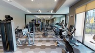 Located at 351 Northwood Dr in Flower Mound, Texas, this single-floor fitness center offers a well-appointed and thoughtfully designed space for a comprehensive workout experience. The main gym area is outfitted with a wide selection of cardio machines — including treadmills, ellipticals, and stationary bikes — alongside a robust lineup of strength and resistance training equipment. A distinctive hexagonal-patterned carpet tiles the floor, adding a modern geometric aesthetic throughout the space. Floor-to-ceiling mirrors along one wall amplify the sense of openness, while recessed and pendant lighting ensures the area is bright and functional. A built-in water station with a mosaic tile accent wall and a coffee station provide convenient amenities between workouts. A separate group exercise studio connects to the main floor through glass-paneled doors. This open, airy room features smooth hardwood-style flooring, a tray ceiling with warm ambient lighting, large roller-shade windows that invite natural light, and full-length mirrored walls. The studio is stocked with stability balls, medicine balls, resistance tools, and yoga mats, making it ideal for classes or individual training sessions. Together, the two areas create a versatile, modern fitness environment designed to support a full range of exercise activities.