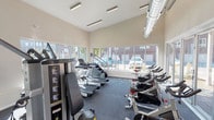 This single-floor fitness center is thoughtfully designed to support a comprehensive workout experience in a bright, open environment. The space features soaring vaulted ceilings that create an airy, expansive atmosphere, complemented by large floor-to-ceiling windows and glass panel doors that flood the room with natural light and offer views of the surrounding outdoor areas, including a landscaped courtyard and patio.

The cardio section is well-appointed with multiple Spirit treadmills, stair climbers, and a row of spin bikes positioned near the glass doors for an energizing, open-air feel. Strength training needs are addressed by a Freemotion multi-function cable machine and a dumbbell rack, providing a versatile range of resistance options. The flooring throughout is durable dark carpet, suitable for high-traffic exercise use.

Overhead, exposed ductwork and linear LED lighting fixtures contribute to the modern, functional aesthetic. A wall-mounted dual water fountain station offers convenient hydration for users. Large windows on multiple walls ensure cross-ventilation and continuous views of the outdoors, enhancing the workout environment. The glass folding doors open directly to the outdoor patio area, seamlessly blending indoor and outdoor fitness spaces.