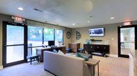 This single-floor space functions as a combined lounge and workspace, thoughtfully designed to accommodate both relaxation and productivity. Large windows along one wall flood the interior with natural light and offer pleasant views of surrounding greenery, creating an inviting, open atmosphere. A patterned valance with a geometric design frames the windows, adding a decorative touch to the space.

The lounge area features a neutral-toned sofa accented with teal and patterned throw pillows, arranged around a low wooden coffee table, providing a comfortable setting for casual gatherings. A wall-mounted flat-screen television is positioned above a dark wood media console, making the area suitable for entertainment and communal viewing.

On the opposite side, a dedicated workspace includes a desk with a desktop computer setup, complemented by dark wood storage furniture and blue accent stools. Decorative wall art and sculptural pieces add visual interest throughout.

The textured ceiling with recessed lighting ensures consistent illumination across the room, while light wood-style flooring ties the contemporary furnishings together. Overall, the space balances functional amenities with a welcoming, modern aesthetic.