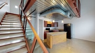 This two-story loft at Artek Lofts showcases a distinctive industrial aesthetic blended with modern finishes throughout. The main level features an open-plan kitchen equipped with rich cherry wood cabinetry, granite-topped center island, black appliances including a refrigerator, dishwasher, and range, all set beneath exposed metal deck ceilings with visible ductwork that reinforces the industrial character of the space. Polished terrazzo-style concrete floors extend throughout both levels, providing a durable and visually striking foundation. A striking open staircase with a bold red steel frame and glass panel railings connects the two floors, serving as both a functional and architectural centerpiece. The upper level features a glass and wood-framed mezzanine railing that overlooks the lower floor, while soaring ceilings with exposed steel trusses and metal deck panels amplify the sense of volume and openness. Multiple windows on both levels allow natural light to filter in, brightening the otherwise industrial palette of white walls and raw materials. The bathroom is finished in clean white subway tile and square floor tile, with a pedestal sink and bathtub/shower combination. Additional rooms on both levels offer flexible, open spaces ideal for a variety of uses.