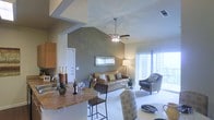 This single-floor residence showcases an open-concept layout that integrates the kitchen, dining, and living areas into one cohesive, functional space. The most striking architectural feature is the vaulted ceiling in the living area, which rises dramatically and adds a strong sense of volume and airiness to the interior. A ceiling fan is centrally mounted, providing both practical ventilation and a visual anchor to the space.

The kitchen is positioned at the front of the open plan and includes a raised breakfast bar with bar seating, offering a natural division between the cooking and living zones without sacrificing visual connectivity. Warm wood cabinetry and a tile backsplash contribute to the kitchen's inviting character.

The living area is finished with neutral-toned carpet and features warm, earthy wall colors that complement the natural light pouring in through large sliding glass doors. These doors lead to an outdoor patio or balcony, enhancing the sense of openness and providing seamless indoor-outdoor flow. Vertical blinds frame the glass doors, offering light control and privacy.

Overall, the space presents a warm, well-lit, and practical layout suited for everyday living.