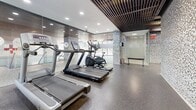 Located at 45 Balliol St in Toronto's Davisville Village neighbourhood, this fitness centre offers a thoughtfully designed workout space spread across a single floor. The facility is defined by its striking interior finishes, including mosaic-tiled accent walls in neutral tones of white, grey, and brown that lend a contemporary, upscale character to the space. Slatted wood ceiling panels add warmth and texture, complemented by recessed pot lighting that ensures the area is consistently well-lit throughout.

The fitness floor is divided into functional zones, each equipped to support a well-rounded workout routine. A dedicated cardio area houses treadmills and an elliptical trainer, positioned alongside floor-to-ceiling mirrored walls that enhance the sense of openness. A strength training section features a full rack of dumbbells, adjustable benches, and multi-station cable machines, all set on durable rubber flooring for safety and comfort. Additional benches and weight equipment are distributed throughout to accommodate a variety of exercise needs.

Glass-panelled dividers and a separate enclosed room provide distinct workout zones within the space. The overall design balances practicality with modern aesthetics, creating a clean and motivating environment for residents to pursue their fitness goals.