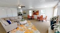 Located at 700 N Albert Pike Ave in Fort Smith, Arkansas, this single-floor community space is designed to bring people together in a welcoming and well-appointed environment. The open floor plan seamlessly connects a comfortable lounge area with multiple dining seating clusters, creating a versatile layout suited for both casual gatherings and organized events. The lounge area features a sectional sofa, a decorative ottoman coffee table, and a bold, colorful area rug that anchors the space with vibrant orange and grey tones. A ceiling fan with integrated lighting provides both comfort and illumination overhead. Two distinct dining areas are furnished with round tables and upholstered chairs in warm coral and salmon hues, complemented by a large floral canvas artwork that enlivens the wall. Natural light streams in through multiple windows along the perimeter, creating a bright and airy atmosphere throughout. Recessed lighting and a skylight further enhance the well-lit interior. In the background, a kitchenette with counter seating offers a functional space for meal preparation or serving. The neutral carpeted flooring ties the entire space together, contributing to the warm and inviting ambiance of this thoughtfully designed community room.