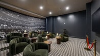 Located at 3111 W Allegheny Ave in Philadelphia, PA, this thoughtfully designed lounge space exudes a sophisticated, cinematic ambiance. The single-floor layout offers a comfortable and intimate gathering environment, making it well-suited for social or entertainment use. Deep navy-blue walls set a moody, upscale tone throughout, complemented by recessed ceiling lighting that casts a warm, focused glow across the room. A bold black-and-white photographic mural of a historic theater audience serves as a dramatic focal point on one wall, reinforcing the venue's entertainment-inspired aesthetic. Opposite the mural, a large wall-mounted flat-screen display is framed by classic red velvet rope stanchions, adding a playful nod to traditional cinema and event spaces. The floor is finished with a distinctive houndstooth-patterned carpet in black and white, grounding the design with a timeless, graphic element. Seating throughout the space consists of richly upholstered deep-green velvet club chairs arranged in clusters around round wooden side tables, each accented with elegant table lamps featuring warm gold bases. The overall atmosphere balances retro-theatrical charm with modern lounge sensibility, creating an inviting and visually cohesive environment ideal for gatherings, screenings, or private events.