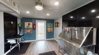 Located at 5757 Wooldridge Rd in Corpus Christi, Texas, this single-floor pet grooming studio is purpose-built for professional animal care. The space combines functional design with a welcoming atmosphere, making it well-suited for both groomers and their four-legged clientele.

The interior is finished with light wood-look flooring and a mix of teal shiplap and white paneled walls, giving the studio a clean, polished look. Recessed lighting and a central ceiling fan with a drum pendant light keep the space bright and comfortable. Charming, colorful dog portrait artwork is displayed throughout, adding personality and warmth to the environment.

The centerpiece of the studio is a large stainless steel professional grooming tub, complete with a built-in ramp for easy pet access and a hose attachment for bathing. Positioned against a striking black diamond-plate panel wall, the tub area is both practical and visually distinctive. Adjacent to the tub is a dedicated grooming table outfitted with a grooming arm, providing a complete workstation for finishing services.

Tools and supplies are neatly organized on pegboard storage nearby, keeping essentials within easy reach. A glass-paneled entry door allows natural light to filter in while offering a view of the outdoor surroundings. The studio is also equipped with a fire extinguisher and climate control, ensuring safety and comfort. Anti-fatigue mats are placed at key work areas throughout the space.