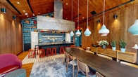Located at Greyrock Place in Stamford, Connecticut, this thoughtfully designed single-floor space makes a bold architectural statement with its dramatic vaulted ceiling clad entirely in warm wood paneling, creating a chalet-like atmosphere that feels both expansive and inviting. Rich hardwood flooring runs throughout, complementing the wood-wrapped walls and ceiling for a cohesive, nature-inspired aesthetic.

The open-plan layout integrates several functional zones seamlessly. A fully equipped kitchen anchors one end of the space, featuring a large central island topped with a striking stainless steel range hood, teal tile backsplash, open blue-painted shelving stocked with colorful dishware, and a retro-style refrigerator. Red bar stools line the island, adding a vibrant pop of color.

A casual lounge area sits adjacent, furnished with eclectic seating and a floor-to-ceiling teal built-in bookcase filled with books and lush potted plants. A long communal dining table with pendant lighting occupies the center of the space, ideal for group gatherings or meetings.

Floor-to-ceiling glazed walls and doors at one end flood the interior with natural light and offer direct views of the surrounding wooded outdoor area, where an exterior patio is visible. Wall-mounted spotlights and pendant fixtures provide warm, layered illumination throughout.
