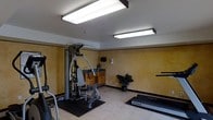 This single-level fitness room offers a well-equipped workout space designed to support a range of exercise routines. The room features warm yellow textured walls accented by a dark trim border, giving the space a distinct and inviting character. Overhead, rectangular fluorescent light fixtures provide bright, even illumination throughout the area.

The fitness equipment includes a multi-station strength training machine, a True-brand treadmill, and an elliptical trainer, all positioned on protective rubber floor mats atop light-toned tile flooring. The layout is thoughtfully organized to allow reasonable circulation between machines.

Along one wall, a built-in cabinetry unit with a dark countertop and an integrated sink adds a practical convenience element, ideal for quick cleanups or storing fitness accessories. A wall-mounted device panel, likely a thermostat or sound system control, is positioned nearby. A decorative potted plant near the cabinetry introduces a touch of greenery, softening the utilitarian nature of the space. Overall, the room presents a functional and neatly maintained environment suited for regular fitness use.
