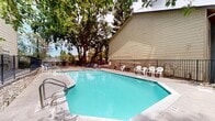 Located at 5341 Walnut Ave in Sacramento, California, this outdoor pool area offers a well-maintained communal retreat designed for relaxation and recreation. The centerpiece is a generously sized, geometrically shaped swimming pool with clear turquoise water, accessible via stainless steel entry rails on multiple sides. The pool's distinctive angular design maximizes usable swimming space while fitting naturally within the enclosed outdoor setting.

The surrounding deck is finished in a smooth concrete surface, providing ample space for lounging. White plastic chairs are arranged along the poolside, offering convenient seating for residents and guests. Additional seating and picnic table arrangements are visible at the perimeter, extending the social potential of the space.

The pool area is enclosed by a black wrought-iron security fence, ensuring safety and privacy. Utility panels are mounted along the building exterior, keeping pool systems organized and accessible. The adjacent building features a light yellow wood-panel siding with a pitched roofline, contributing to the residential character of the setting.

Mature trees frame the space on multiple sides, providing natural shade and a lush, green backdrop that softens the built environment. The combination of open sky, tree canopy, and well-kept water creates an inviting outdoor atmosphere well-suited to Sacramento's warm climate.