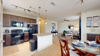 Located at 3010 W Loop 1604 N Access Rd in San Antonio, Texas, this single-story residence offers a thoughtfully designed open-concept layout that seamlessly connects the kitchen, dining, and living areas. The kitchen is a standout feature, showcasing rich dark-wood cabinetry, granite countertops, a central island with pendant lighting, tile flooring, and black appliances including a French-door refrigerator. Track lighting and a window above the sink bring additional brightness to the space. The dining area sits adjacent to the kitchen and is bathed in natural light from large windows dressed with navy blue curtains and horizontal blinds, creating a warm and inviting atmosphere. The living room continues this bright, airy feel with plush carpeting, a ceiling fan, and generous window views of the surrounding greenery. Both bedrooms are carpeted and well-lit with large windows and ceiling fans, offering comfortable and restful retreats. The bathroom features tile flooring, a granite-topped dark-wood vanity, and a soaking tub set within an elegantly arched alcove with tile surround. A dedicated in-unit laundry area with full-size washer and dryer and built-in shelving adds everyday convenience. Throughout, the space maintains a cohesive, neutral palette with warm finishes.