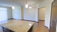 This single-floor space is designed with an open-concept layout that integrates the kitchen and living areas into one cohesive environment. The kitchen is anchored by a generously sized island topped with speckled granite countertops, offering both workspace and casual seating potential. Three pendant lights hang above — two cylindrical white fixtures and one wider bowl-shaped pendant — providing focused task lighting while contributing to the overall aesthetic.

The flooring throughout is a warm hardwood-style surface that runs continuously from the kitchen into the adjoining living area, reinforcing the sense of openness and visual flow. Neutral, light-toned walls complement the natural warmth of the floors and granite, creating a clean and inviting atmosphere.

A wide open passage connects the main living space to an additional room visible to the right, suggesting a flexible layout adaptable to various uses. Dark baseboards provide a subtle contrast against the light walls, adding a finished, polished detail. The overall ambiance is bright, understated, and functional, well-suited for everyday living.