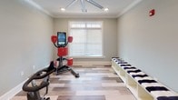 Located at 9001 High Peak Dr. in Greenville, South Carolina, this single-floor fitness studio is thoughtfully designed to support a well-rounded exercise routine. The space features warm-toned hardwood-style flooring laid in a varied plank pattern, crisp white crown molding, and soft sage-green walls that create a clean, energizing atmosphere throughout.

The main workout area is equipped with multiple stationary cycling bikes, ideal for cardio training sessions, and a functional combat training machine with an interactive display screen for guided workouts. A large window with white blinds allows natural light to fill the room, enhancing the open feel of the space. Overhead, a large ceiling fan with recessed lighting ensures comfortable air circulation during intense training sessions.

Along one wall, a row of white cubby-style storage benches with navy and white striped cushioned tops provides convenient seating and storage for personal belongings. A glass-paneled door connects to an adjacent area, allowing for easy flow between spaces.

The studio's combination of practical equipment, polished finishes, and bright, airy design makes it a functional and inviting environment for fitness enthusiasts of all levels.