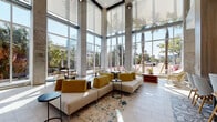 This striking lobby in Jacksonville, Florida exemplifies contemporary design with an open, airy aesthetic anchored by dramatic double-height ceilings and expansive floor-to-ceiling glass walls. Exposed concrete columns frame the space, lending an industrial elegance that complements the polished tile flooring throughout.

The main lounge area is thoughtfully arranged with neutral-toned sectional sofas, mustard-yellow accent chairs, and sleek black side tables, all set atop a vibrant teal and cream area rug. Multiple seating vignettes accommodate both casual conversation and individual use, including white lounge chairs with gold-finished frames positioned directly before panoramic windows that frame views of swaying palm trees and lush landscaping.

A secondary lounge zone features intimate booth-style seating beneath a warm wood slat ceiling detail and soft pendant lighting, offering a more sheltered environment within the larger open plan. An upper-level mezzanine with wood-paneled railings overlooks this area, adding vertical depth.

The elevator corridor transitions seamlessly from the main lounge, featuring recessed linear lighting, stainless steel elevator doors, a decorative credenza with sculptural accents, and curated artwork. The entryway incorporates glass doors and botanical wall art, setting a welcoming tone from arrival.