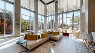 This striking lobby in Jacksonville, Florida exemplifies contemporary design with an open, airy aesthetic anchored by dramatic double-height ceilings and expansive floor-to-ceiling glass walls. Exposed concrete columns frame the space, lending an industrial elegance that complements the polished tile flooring throughout.

The main lounge area is thoughtfully arranged with neutral-toned sectional sofas, mustard-yellow accent chairs, and sleek black side tables, all set atop a vibrant teal and cream area rug. Multiple seating vignettes accommodate both casual conversation and individual use, including white lounge chairs with gold-finished frames positioned directly before panoramic windows that frame views of swaying palm trees and lush landscaping.

A secondary lounge zone features intimate booth-style seating beneath a warm wood slat ceiling detail and soft pendant lighting, offering a more sheltered environment within the larger open plan. An upper-level mezzanine with wood-paneled railings overlooks this area, adding vertical depth.

The elevator corridor transitions seamlessly from the main lounge, featuring recessed linear lighting, stainless steel elevator doors, a decorative credenza with sculptural accents, and curated artwork. The entryway incorporates glass doors and botanical wall art, setting a welcoming tone from arrival.