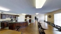 This single-story clubhouse offers a well-designed, open-concept layout that seamlessly combines a fully equipped kitchen with a generous dining area, making it well-suited for community gatherings and events. The kitchen is outfitted with dark wood cabinetry, a large central island with a dark countertop, a white refrigerator with an ice and water dispenser, a white range, and a built-in microwave. Ample cabinet storage lines the walls, providing practical functionality for hosting and catering needs. The hardwood flooring throughout adds warmth and durability to the space. The dining area extends alongside the kitchen and features multiple long tables with seating for a large number of guests, arranged to maximize capacity. A doorway visible from the main area leads to an additional room, suggesting further space for meetings or events. Neutral yellow-toned walls, textured ceilings, and flush-mounted fluorescent lighting contribute to a bright, welcoming atmosphere. A window along the far wall allows natural light to filter into the dining area, enhancing the overall openness of this practical and inviting community space.