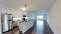 Located at Greybarn Amityville on Broadway in Amityville, NY, this single-floor residence offers a thoughtfully designed open-concept layout with modern finishes throughout. The kitchen is a standout feature, showcasing crisp white shaker-style cabinetry that extends to the ceiling, a classic white subway tile backsplash, dark countertops, and a full suite of stainless steel appliances including a refrigerator, electric range, microwave, and dishwasher. A peninsula with pendant lighting connects the kitchen seamlessly to the expansive living area, which is finished with rich, dark hardwood-style flooring and a large window that fills the space with natural light. The bedroom offers plush carpeting, a clean neutral palette, and ample room for furnishings, with easy access to adjoining spaces. The bathroom is finished with white subway tile surrounding the soaking tub and shower combination, small-format mosaic floor tile, and white cabinetry with dark countertops, delivering a bright and timeless aesthetic. Throughout the residence, white walls, black hardware accents, and consistent flooring create a cohesive, contemporary ambiance that is both functional and inviting.