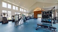 This modern fitness center is designed to accommodate a wide range of workout needs within a single, thoughtfully organized facility. The main exercise floor is defined by a dramatic vaulted ceiling that rises to impressive heights, creating an open, airy atmosphere. Rows of tall windows line the walls, flooding the space with natural light and offering views of the surrounding outdoors. The durable blue rubber flooring extends throughout, providing both safety and a clean, contemporary aesthetic.

The main floor is comprehensively equipped with Matrix-brand strength machines, adjustable benches, a multi-functional cable and free weight station, medicine ball racks, and a full selection of dumbbells. Cardio equipment, including multiple treadmills, ellipticals, cycling bikes, and a stair climber, is thoughtfully distributed throughout the space. A striking wood-panel accent wall serves as an architectural focal point, complemented by large mirrored sections that enhance the sense of depth.

A separate yoga and movement studio adjoins the main floor, offering light hardwood flooring, full-length mirrors, a ballet barre, a large display screen, and an assortment of stability balls, resistance bands, and yoga accessories. The studio features decorative wainscoting and textured wallpaper, giving it a distinct, calming character ideal for stretching, yoga, or group fitness activities.