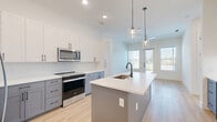 Located at 4220 Tributary Way in Frisco, TX, this well-designed two-bedroom, two-bathroom residence offers a thoughtful single-floor layout with contemporary finishes throughout. The open-concept kitchen and living area serves as the heart of the home, featuring a central island with an undermount sink and matte black faucet, crisp white upper cabinetry, two-tone gray lower cabinets, white quartz countertops, a subway tile backsplash, and stainless steel appliances including a refrigerator, range, and over-the-range microwave. Glass pendant lights add a polished touch above the island. The living area extends naturally from the kitchen, bathed in natural light from large windows and a glass-panel door that opens onto a private balcony with outdoor views. Light hardwood-style flooring runs through the main living areas, while plush carpet adds warmth to the bedrooms. The primary bedroom is a comfortable retreat with direct access to an en-suite bathroom featuring a dual-sink vanity with white cabinetry and matte black hardware, a vaulted ceiling, and a dedicated walk-in closet with built-in shelving and hanging rods. The second bedroom is bright and well-proportioned, with a window overlooking the outdoors. Two closets provide ample storage throughout the home, completing this clean, modern residence.